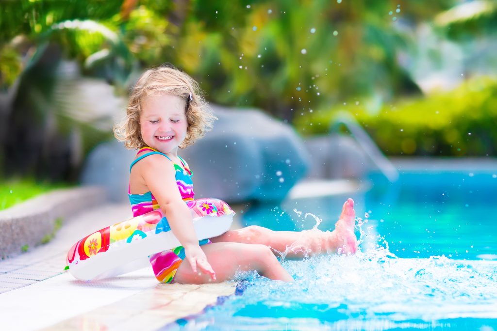 Adorable little girl with curly hair wearing a colorful swimming suit playing with water splashes at beautiful pool in a tropical resort having fun during family summer vacation
